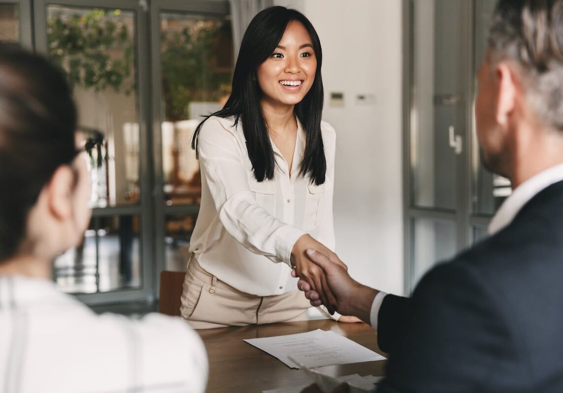 Business, career and placement concept - image from back of two employers sitting in office and shaking hand of young asian woman